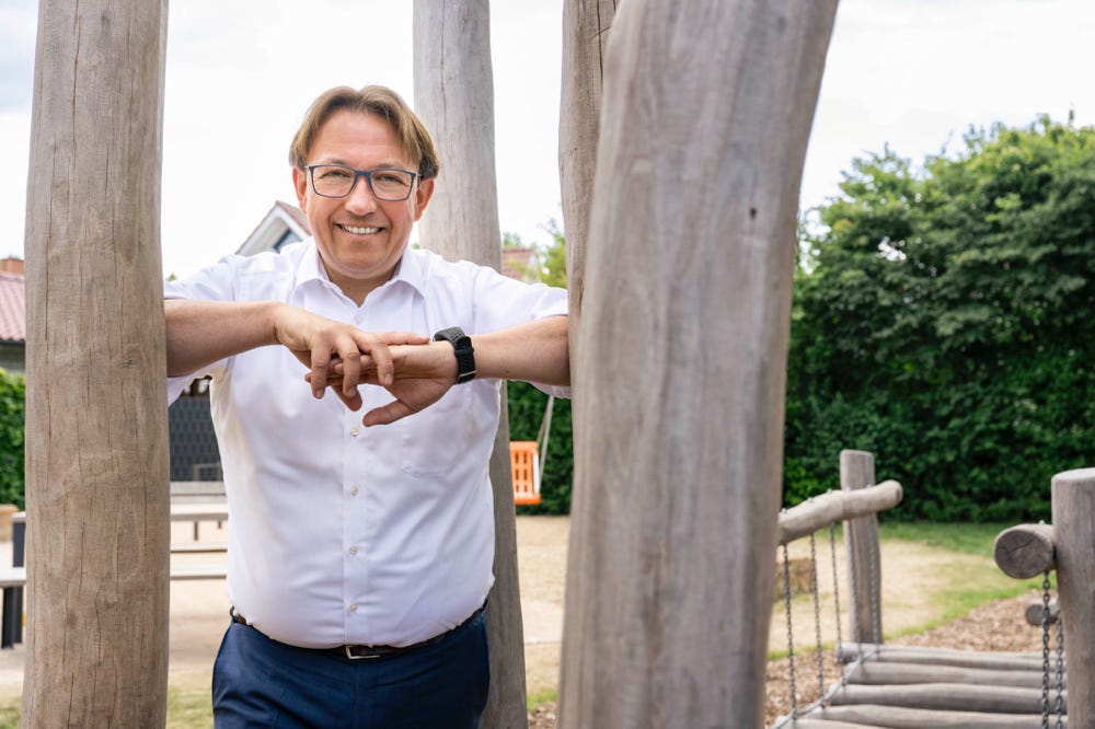 Marc Schrameyer lehnt zwischen zwei Baumstämmen auf einem Spielplatz und lacht in die Kamera. 